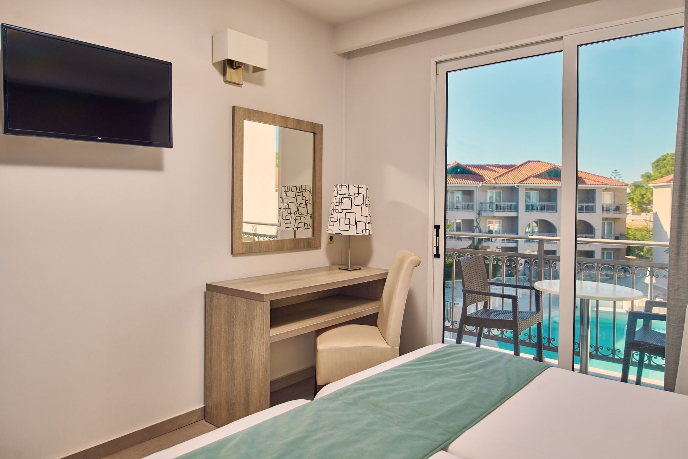 A hotel room with a bed, a desk, a chair, a TV, a mirror, and a balcony with a pool view.