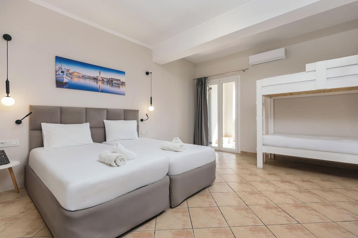 Uma cama de casal com cabeceira, uma mesa de cabeceira com abajur, um quadro com paisagem costeira e uma cama beliche à direita.