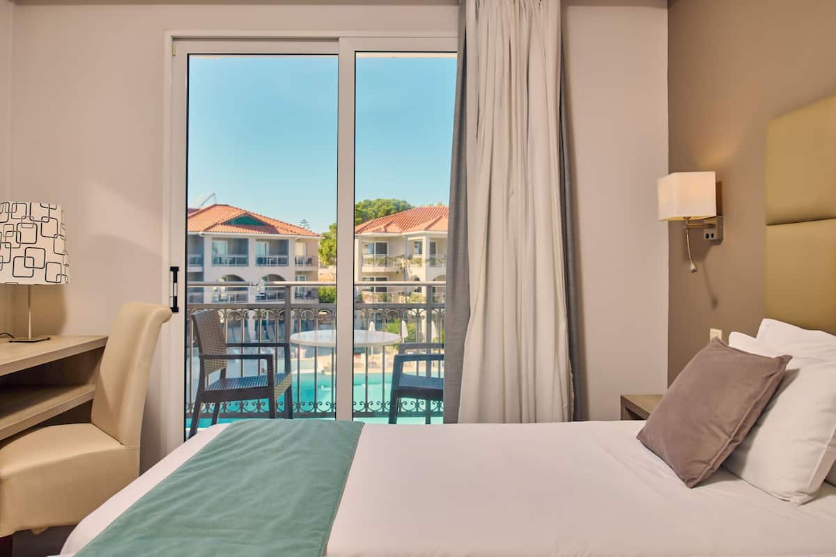Quarto de hotel com cama, escrivaninha, cadeira, mesa de cabeceira, luminária e vista para uma piscina e edifícios.