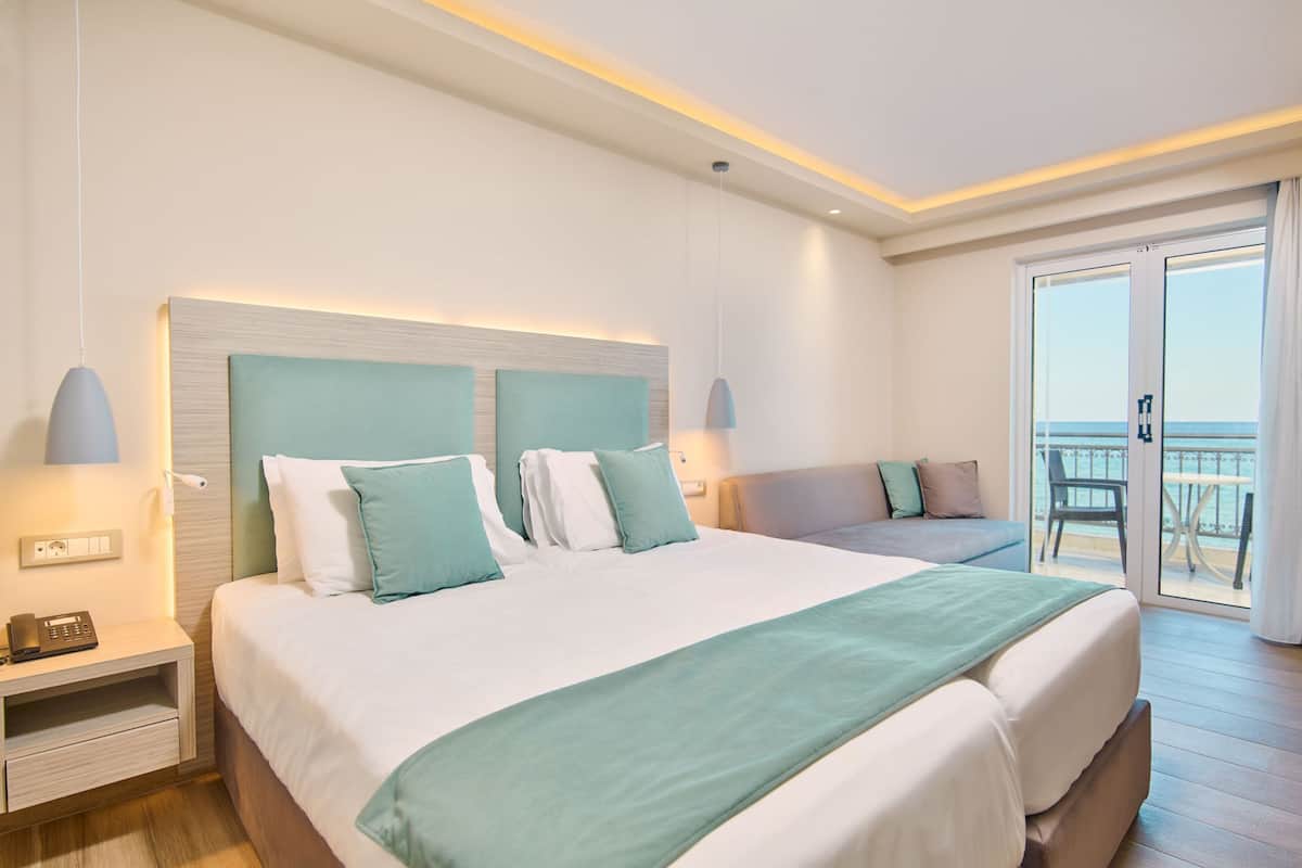 Quarto de hotel com cama grande, mesa de cabeceira com abajur, sofá e varanda com vista.