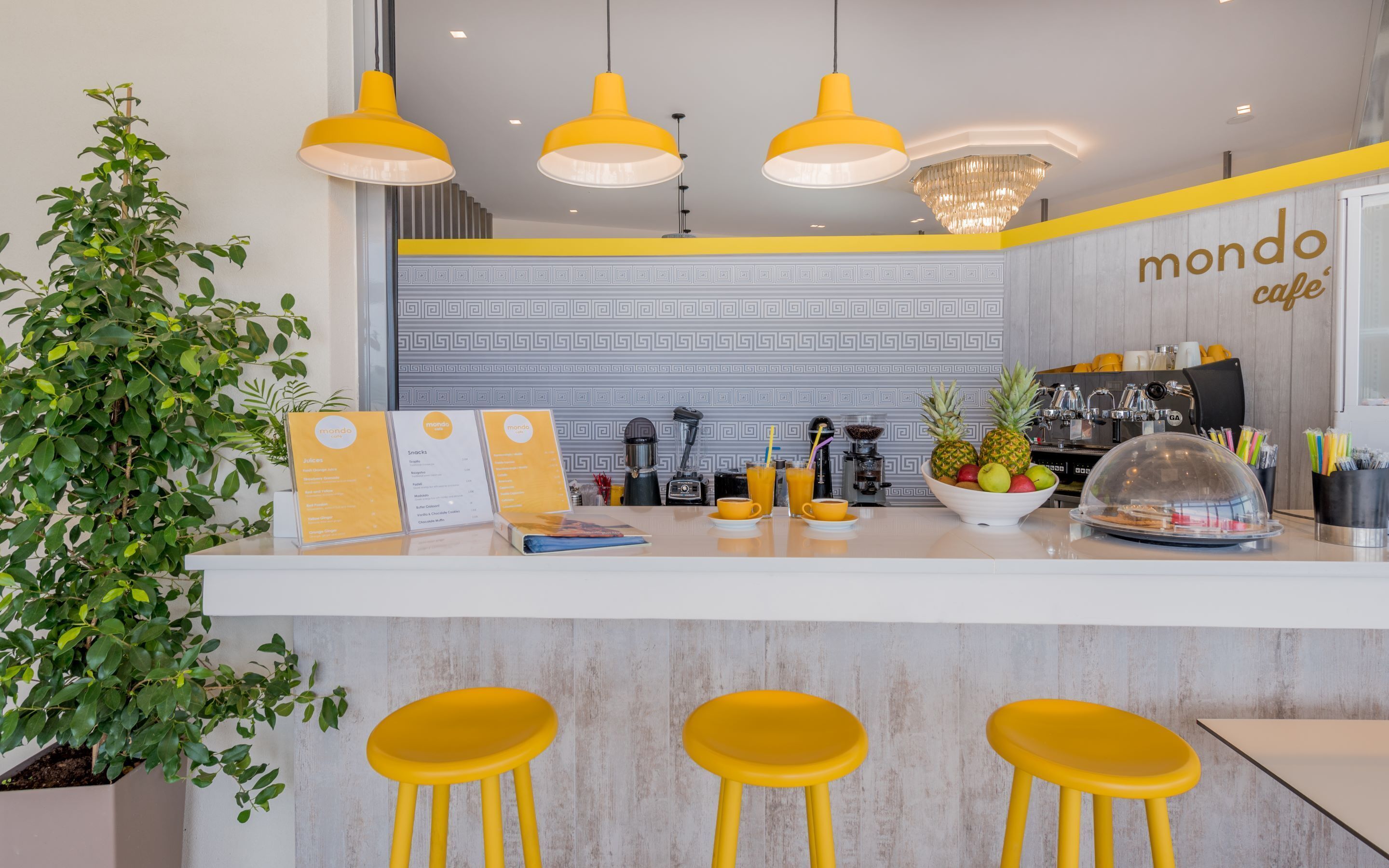 L'intérieur d'un café moderne, avec un bar central, des tabourets jaunes, un panneau affichant le menu et une plante décorative.