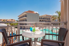 Un balcón con dos sillones de mimbre, una mesa redonda pequeña y dos bebidas. Al fondo, se ve una piscina y un edificio con balcones.