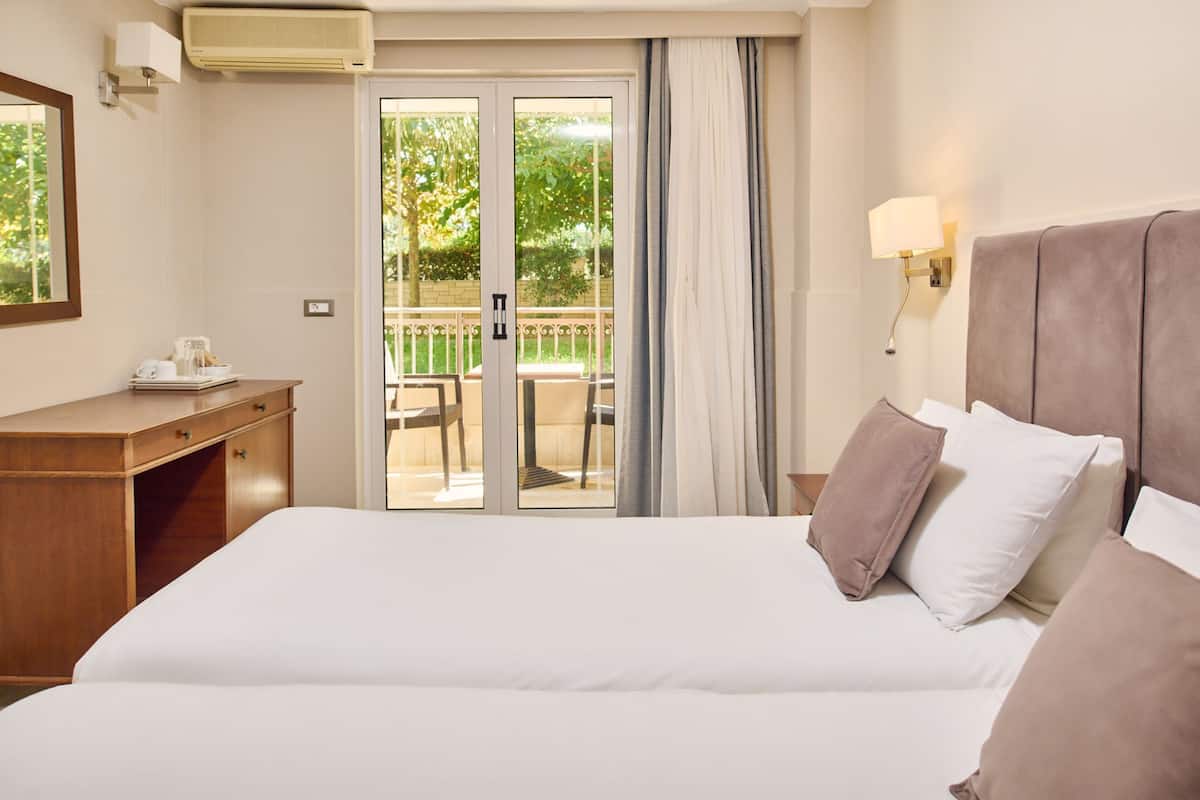 Quarto de hotel com uma cama grande, uma escrivaninha de madeira, uma cadeira e uma varanda com mesa e cadeiras.