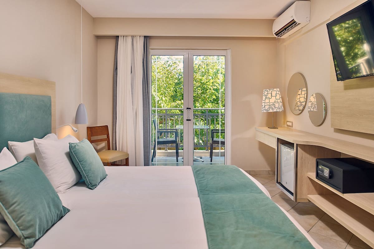 Quarto de hotel com cama, televisão, mini-frigorífico e varanda com uma cadeira.
