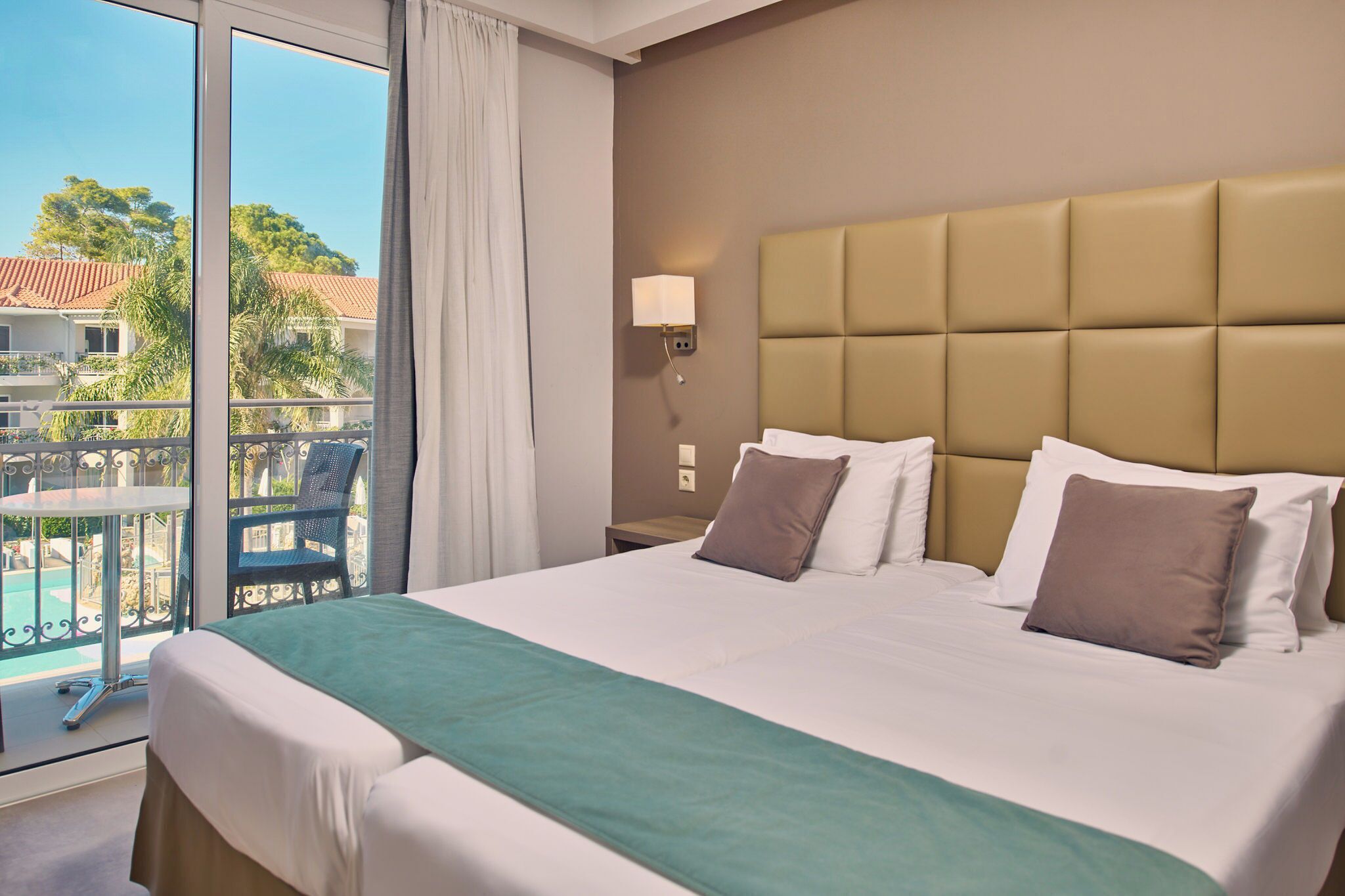 A hotel room with a large bed, a view of a pool and buildings, and a balcony with a chair.