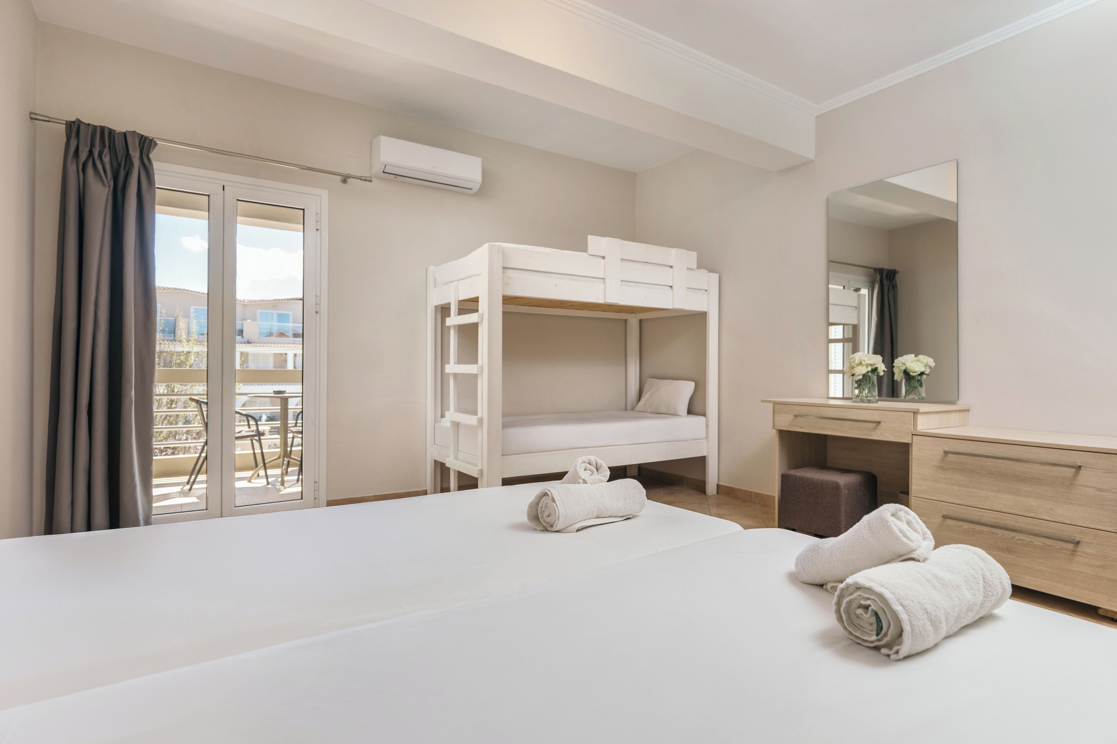 A hotel room with a bunk bed, a large bed, a desk with a mirror, and a view of the outside balcony.