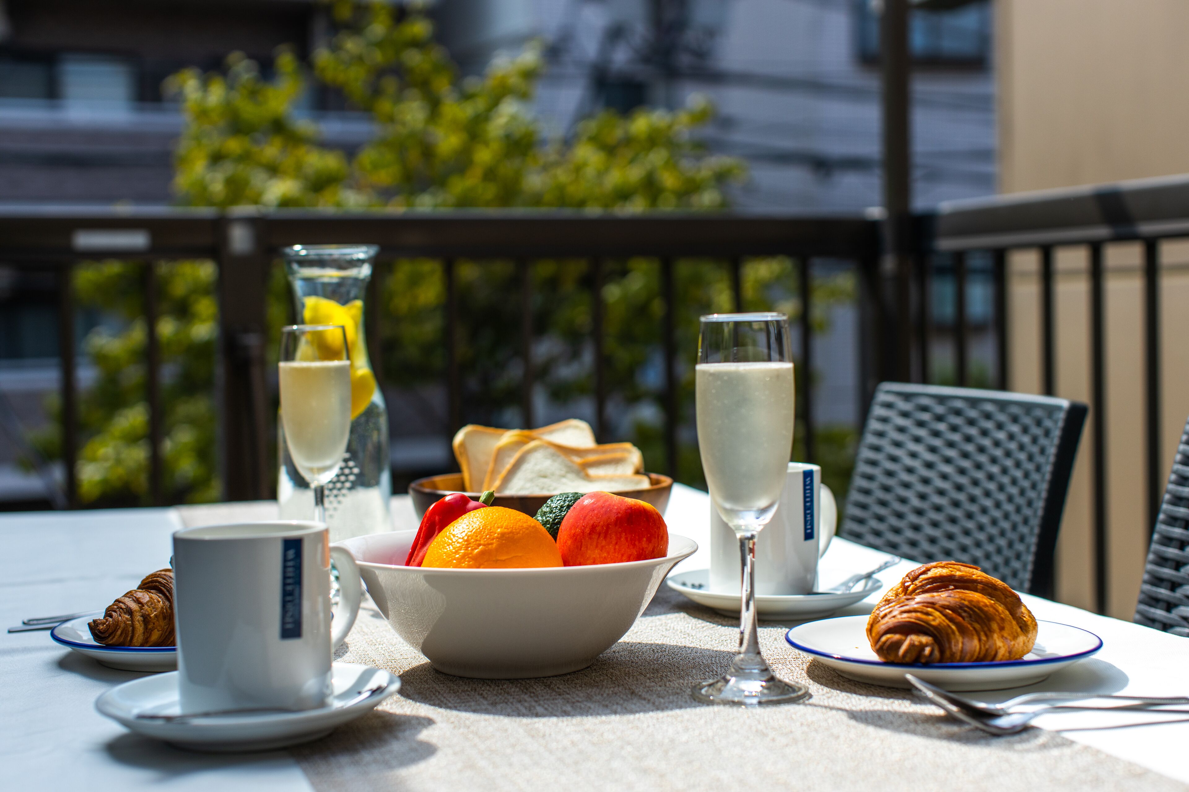 A table set with croissants, a bowl of fruit, a glass of white wine, and a cup of coffee on a balcony with a view of buildings and trees.