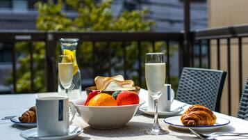 A table set with croissants, a bowl of fruit, a glass of white wine, and a cup of coffee on a balcony with a view of buildings and trees.