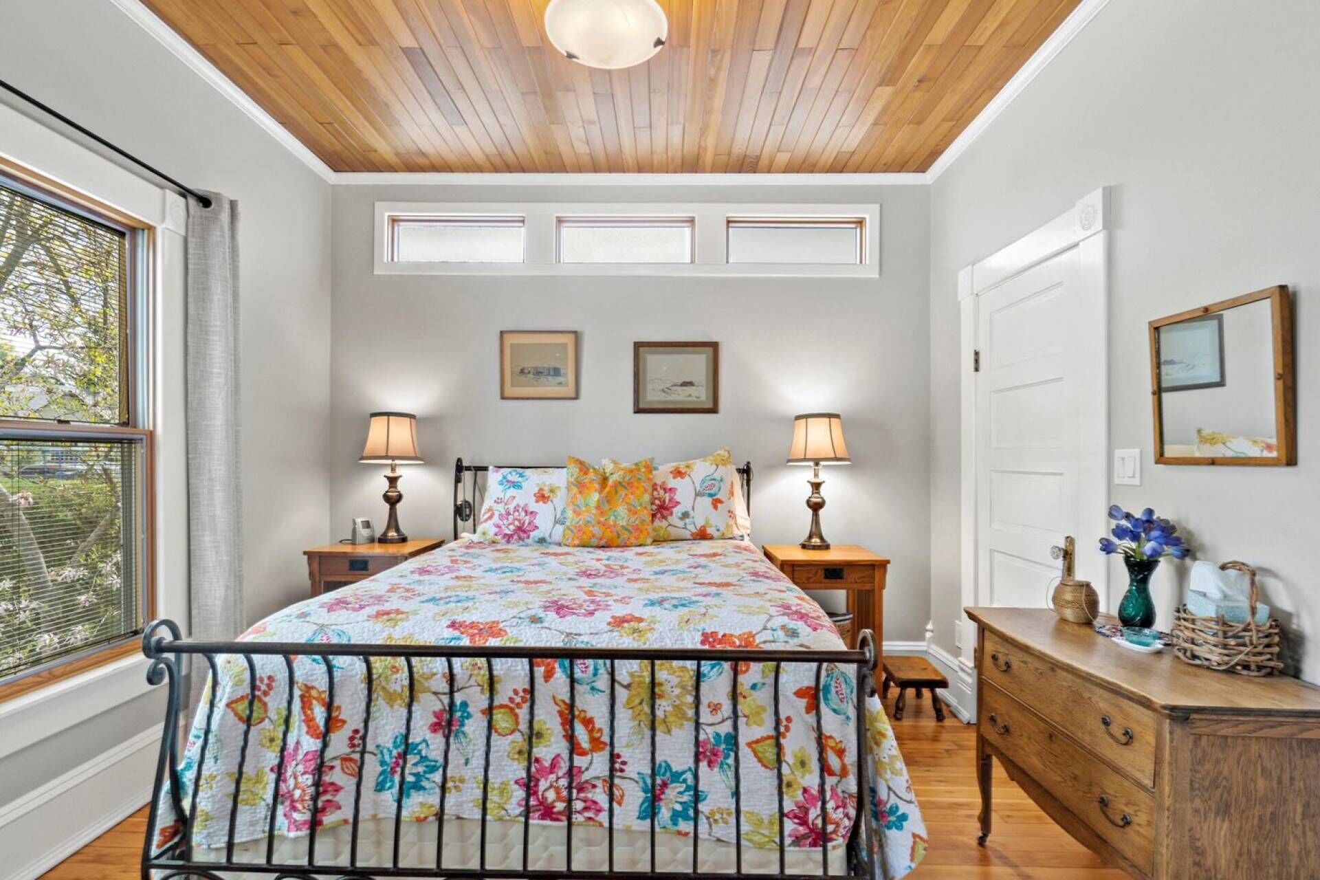 A bedroom with a wooden ceiling, a bed with a floral bedspread, two bedside lamps, a dresser, and a window with blinds.