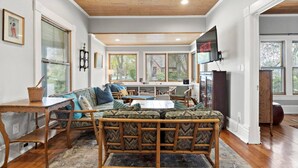 A spacious living room with a large sofa, a coffee table, and a TV mounted on the wall. There are large windows providing natural light and a view of the outdoors. - Cozy North End Cottage! Walk the Tree-lined Neighborhood to Hyde Park + Camel's Back Park (Boise)