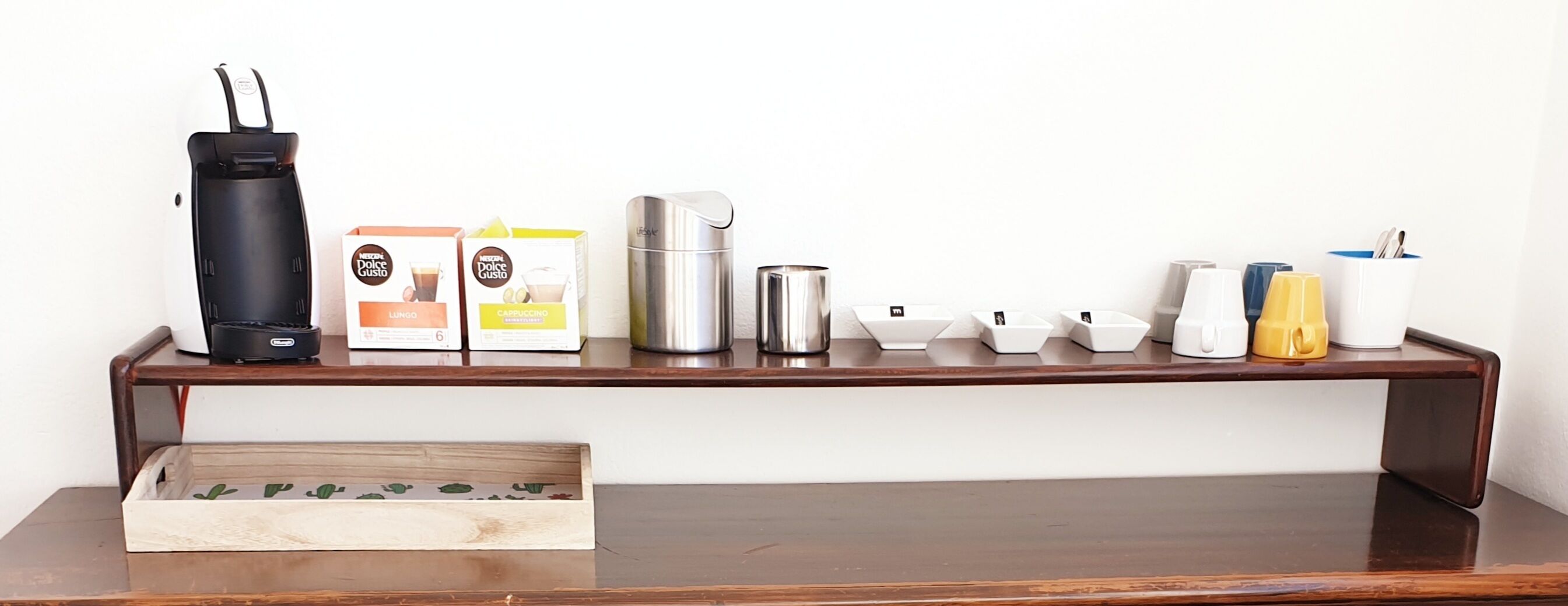 Una cafetera, dos cajas de café Dolce Gusto, un recipiente de acero inoxidable y una bandeja con tazas y un objeto amarillo sobre una mesa de madera.