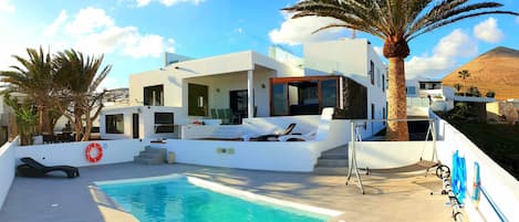 A modern villa with a swimming pool, outdoor seating, and a view of a hill.