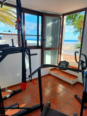 A home gym with exercise equipment, a sliding glass door leading to a balcony with a view of the sea and palm trees.