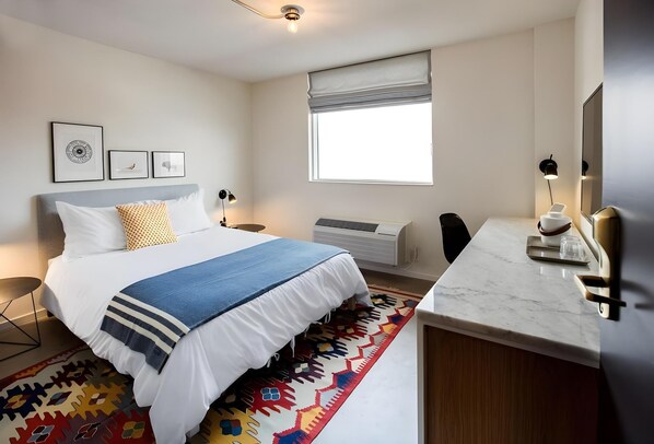 A modern hotel room with a neatly made bed, a large window with blinds, a flat-screen TV, a desk with a lamp, and a patterned rug. - Trumbull and Porter Hotel (Detroit)