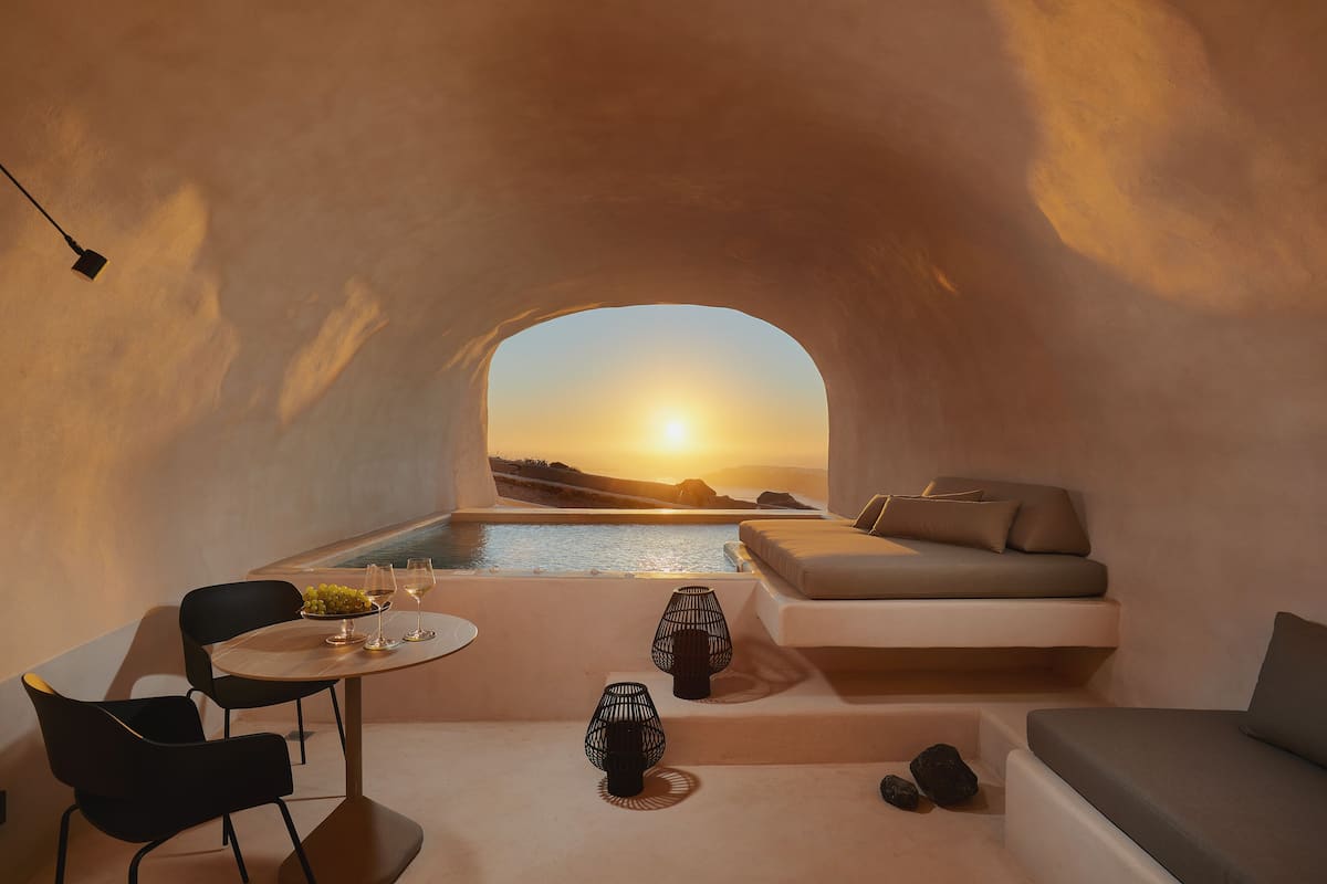 A cozy interior with a seating area, a table with a bowl of fruit and glasses, and a view of the sunset over water.