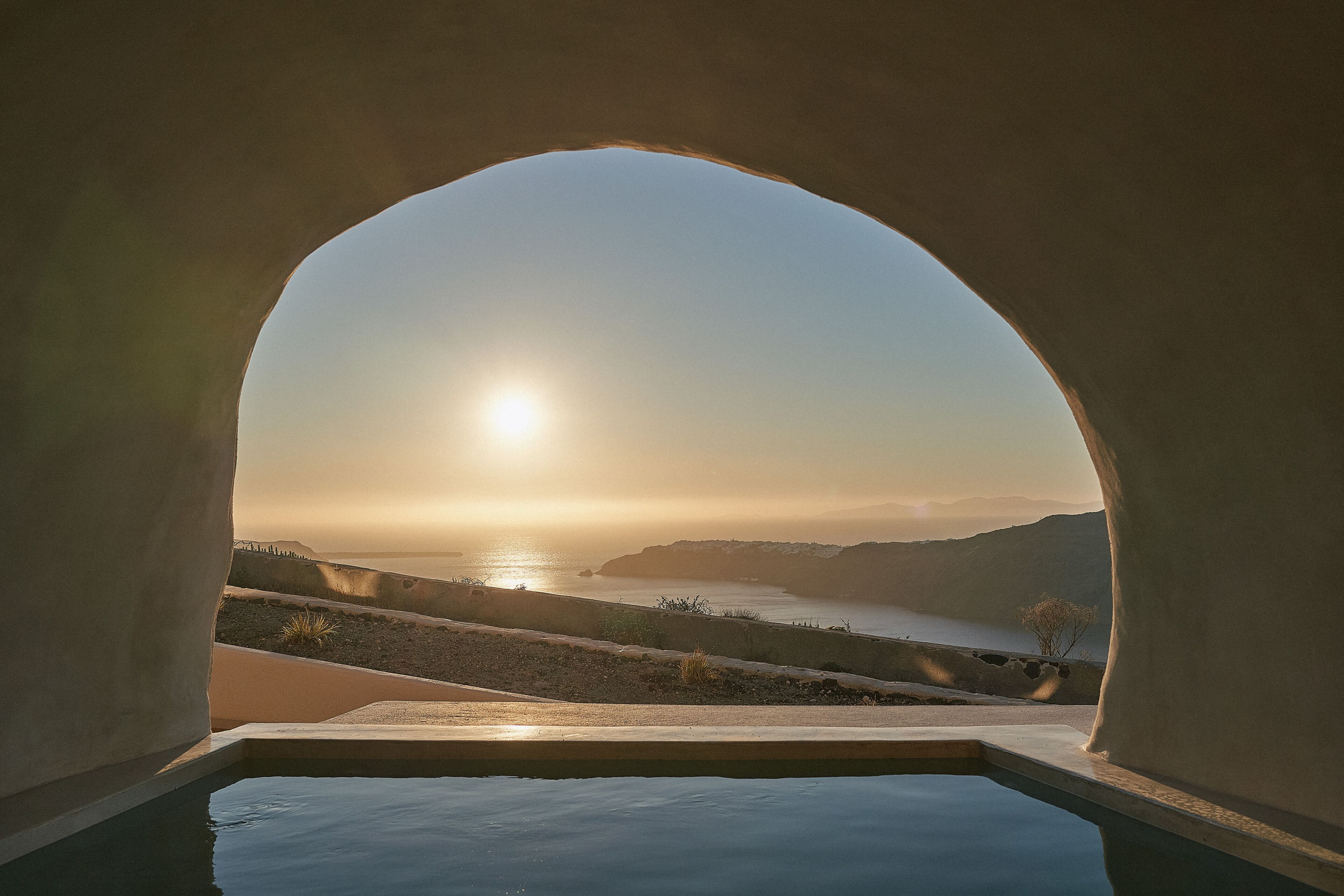 A view through an arched window overlooking a body of water, a hilly landscape, and a clear sky.