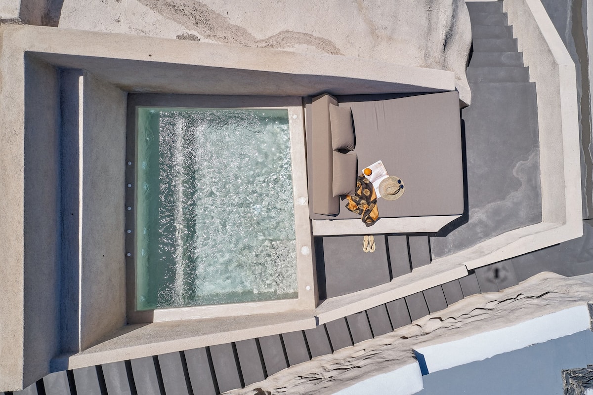 A rooftop pool area with a glass enclosure, a seating area with a cushion, and a small table with a hat and book.