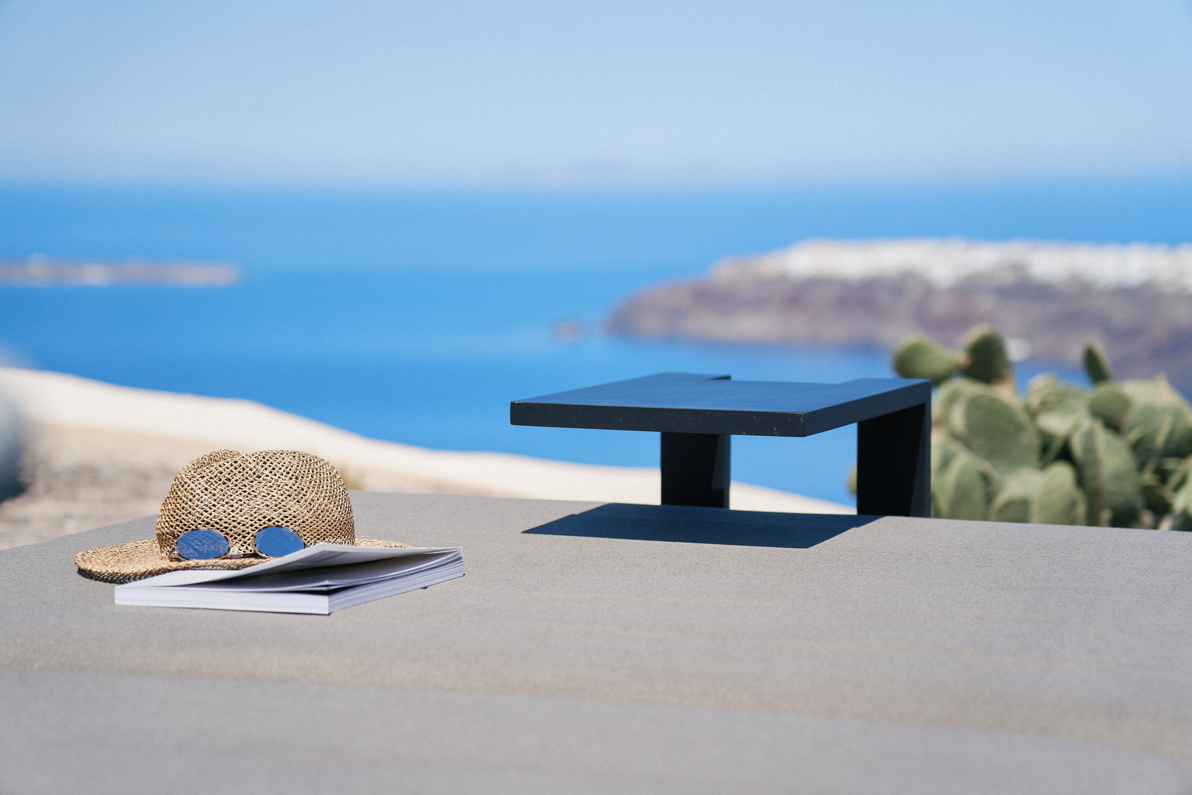 Une table surmontée d’un chapeau et d’un carnet, posée sur une surface en béton, d’où l’on aperçoit une plage avec des cactus et l’océan.