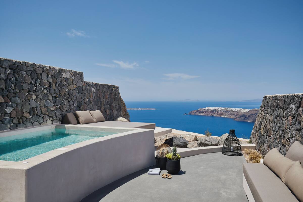 A stone-walled infinity pool with a clear view of the sea, a lounge area with cushions, and a potted plant.