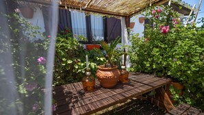 Une table en bois, agrémentée de pots en terre cuite et d’une plante en pot, se trouve dans un coin du jardin, où la végétation luxuriante et les fleurs créent un cadre verdoyant.