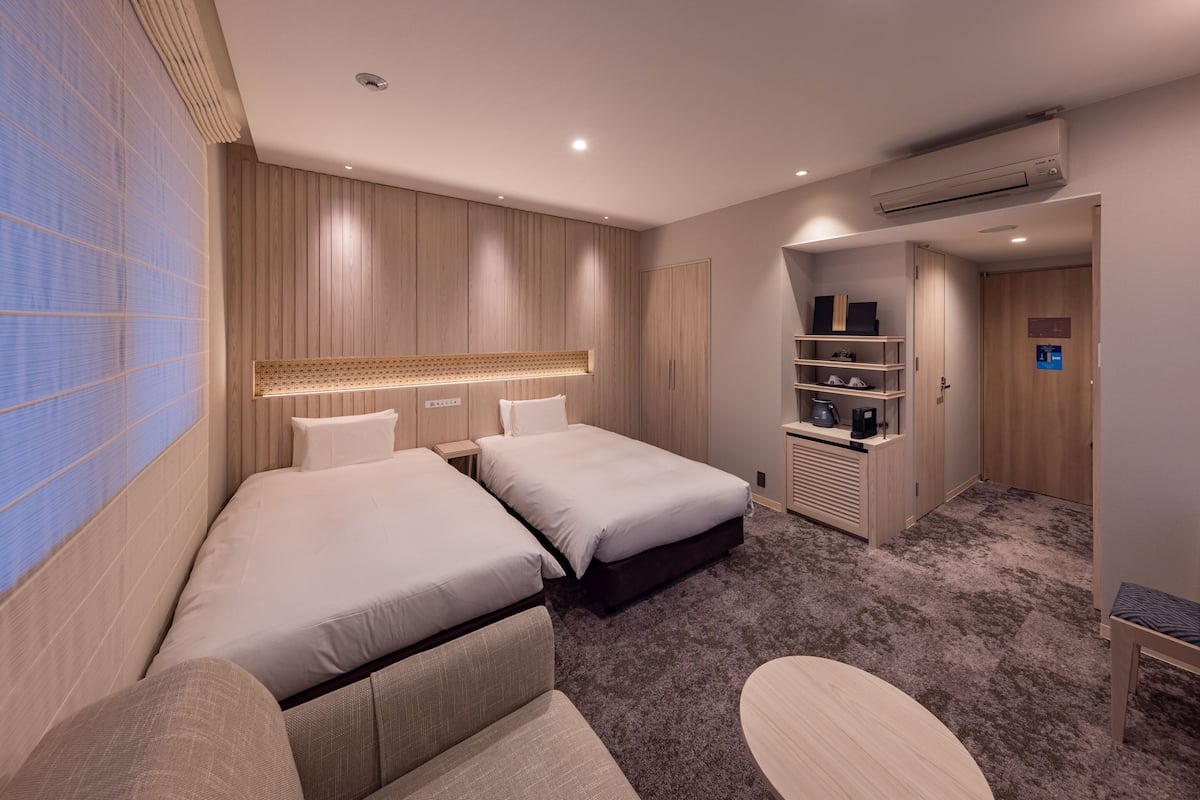 Ein modernes Hotelzimmer mit zwei Betten, einer Schlafcouch, einem kleinen Tisch und einer Küchenzeile.
