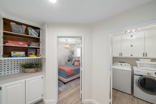 A compact bedroom with a bed, bedside table, and a nightstand. A hallway leads to a bathroom with a washing machine and a laundry basket. - Dune Lake - Completely remodeled and designed for the ultimate relaxing dese... (Bermuda Dunes)
