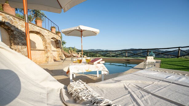 A poolside area with lounge chairs, a table with drinks, and a view of a bridge and hills. - Villa "Il Grillo", privacy in the hills, private pool and panoramic view. (Toscana)