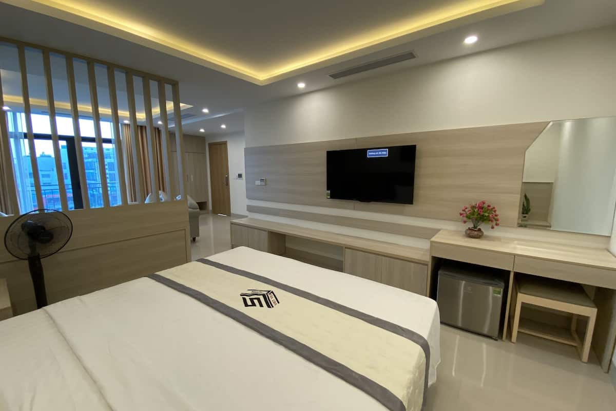 A modern hotel room with a bed, a TV mounted on the wall, a small desk, and a fan.