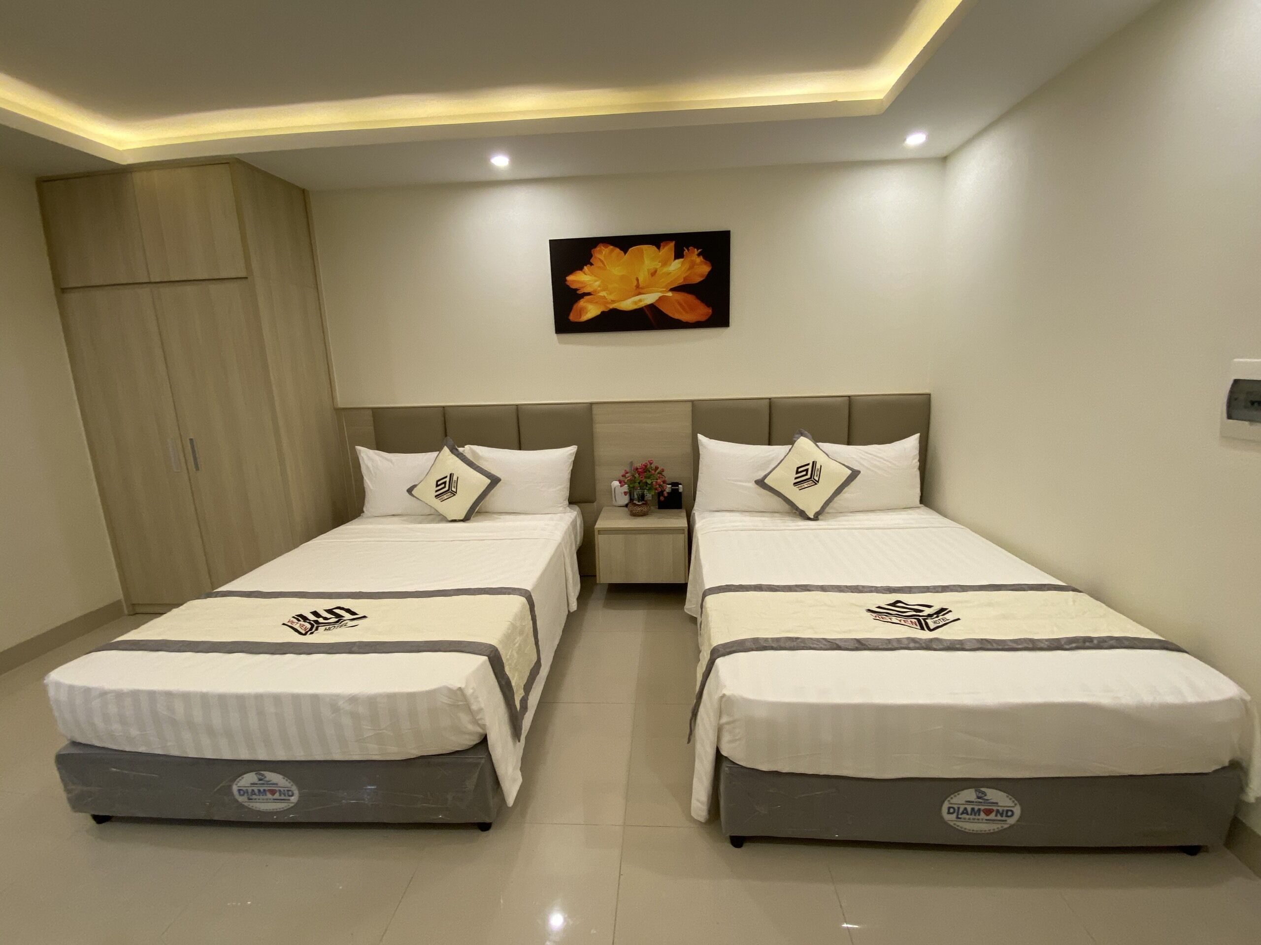 Une chambre d’hôtel avec deux lits, chacun équipé d’un matelas de la marque « Diamond », une table de chevet ornée d’un vase de fleurs et un tableau représentant une fleur accroché au mur.