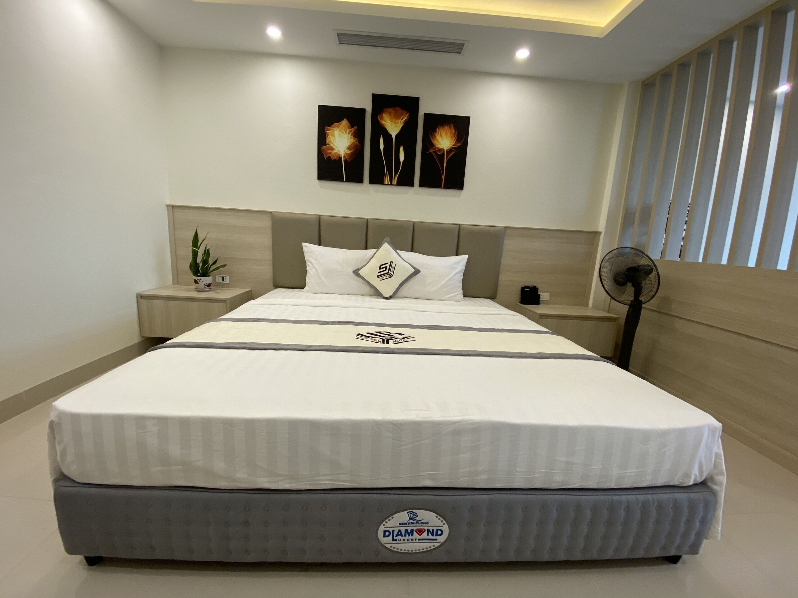 Een moderne hotelkamer met een groot bed, een nachtkastje, een klein tafeltje, een potplant en een ventilator aan de muur.