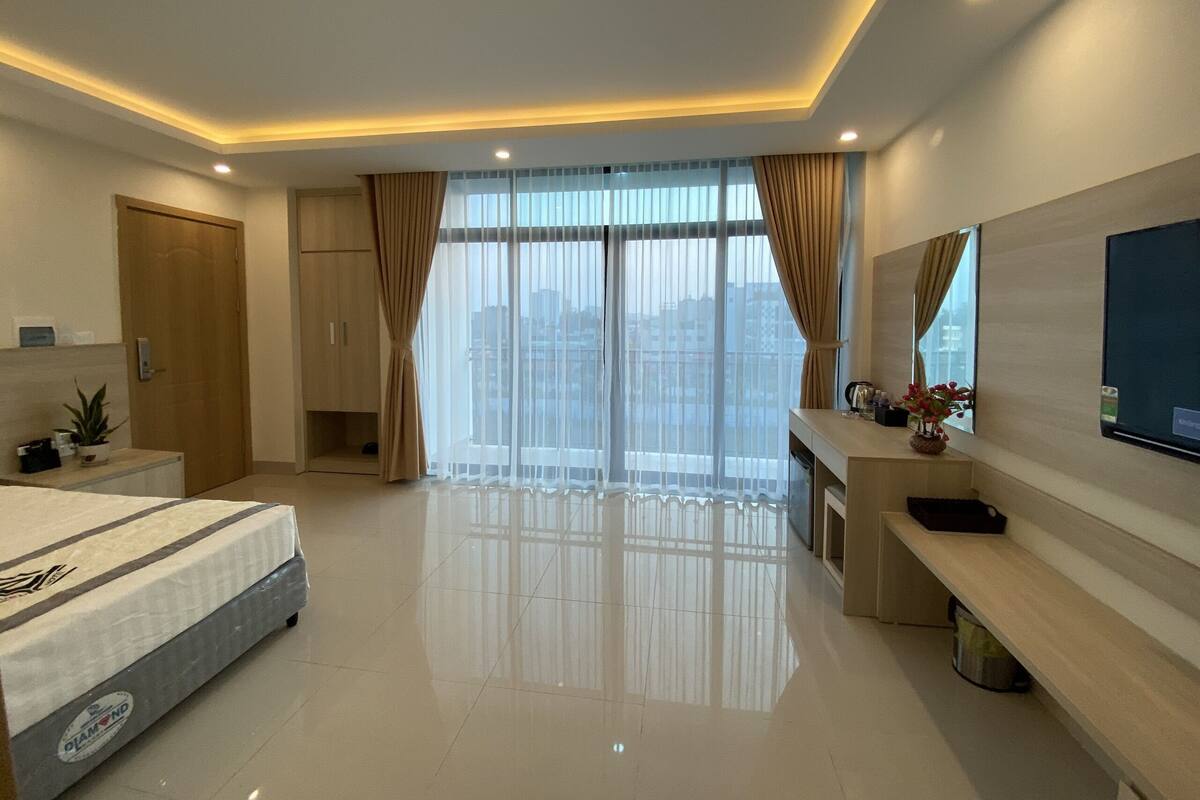 A modern hotel room with a large bed, a desk, a television, and a balcony with curtains.