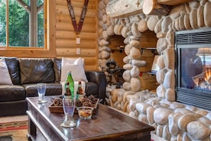 A cozy cabin interior with a stone fireplace, leather sofa, and a wooden coffee table with drinks and snacks.