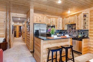 A log cabin interior with a kitchen featuring wooden cabinets, a stainless steel refrigerator, a stove, and a dishwasher. There are two bar stools at the kitchen island.