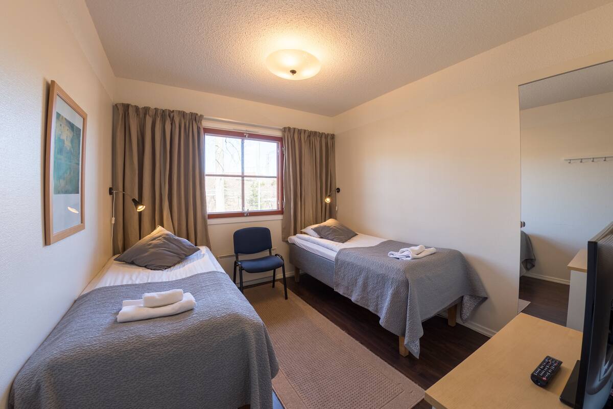 A room with two single beds, a chair, a TV, and a window with curtains.