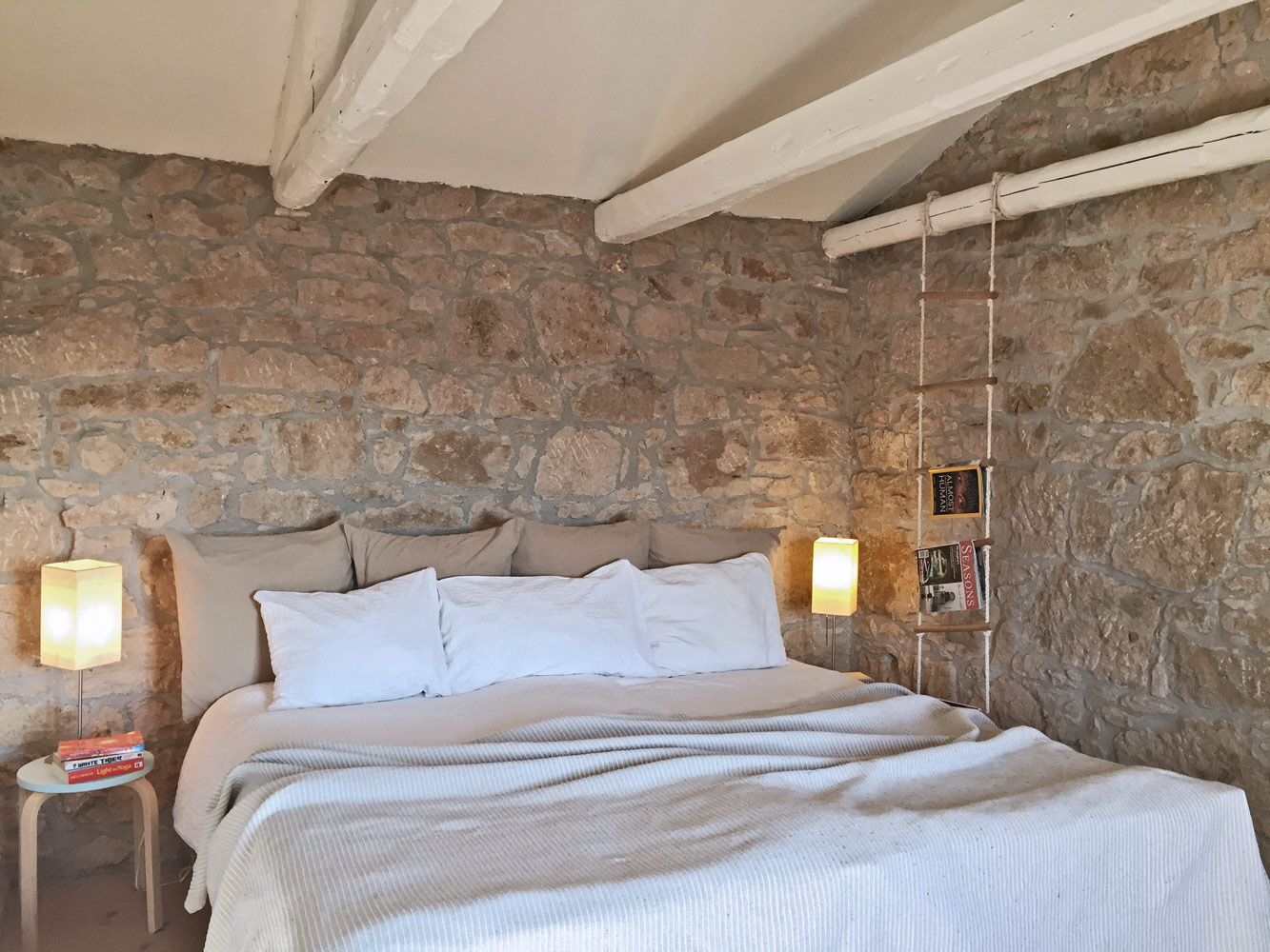 Un dormitorio con pared de piedra, una cama con ropa de cama blanca, dos l ámparas de noche y una mesita con libros.