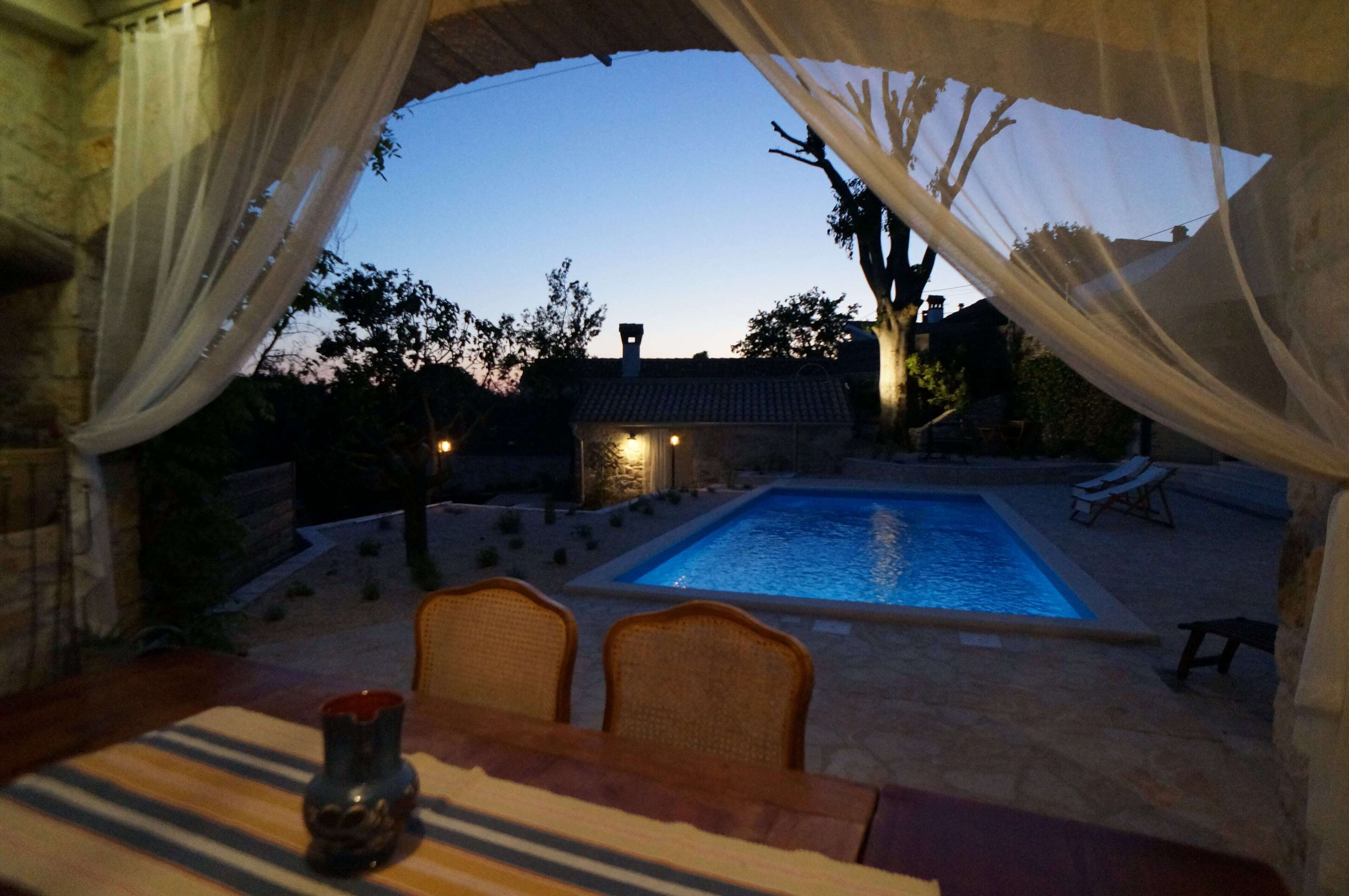 A pool area with a view of a building and trees, surrounded by a stone wall and a covered seating area.