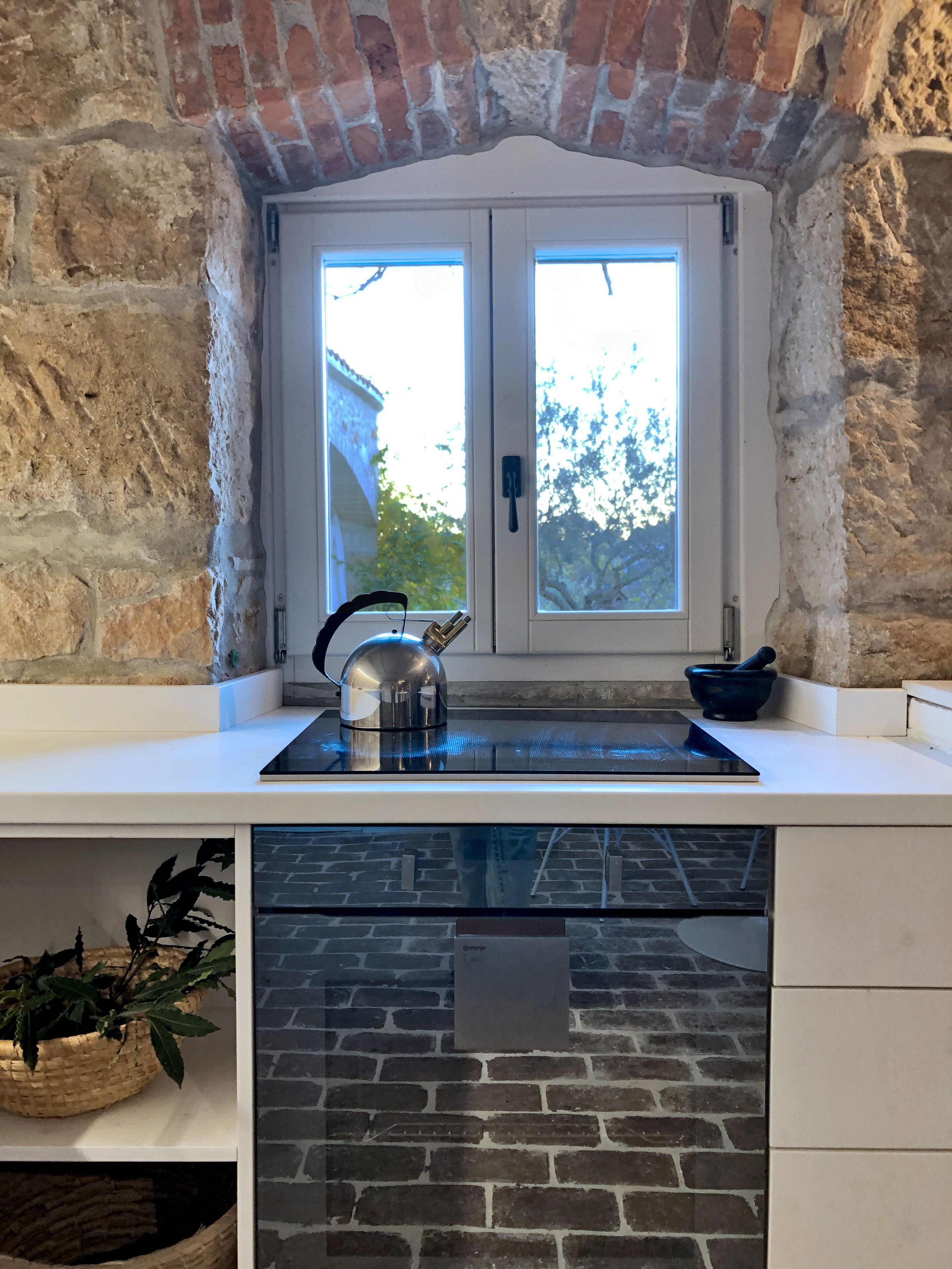 Une cuisine avec un mur en briques, une fenêtre à double porte, une bouilloire, un bol et une plante.