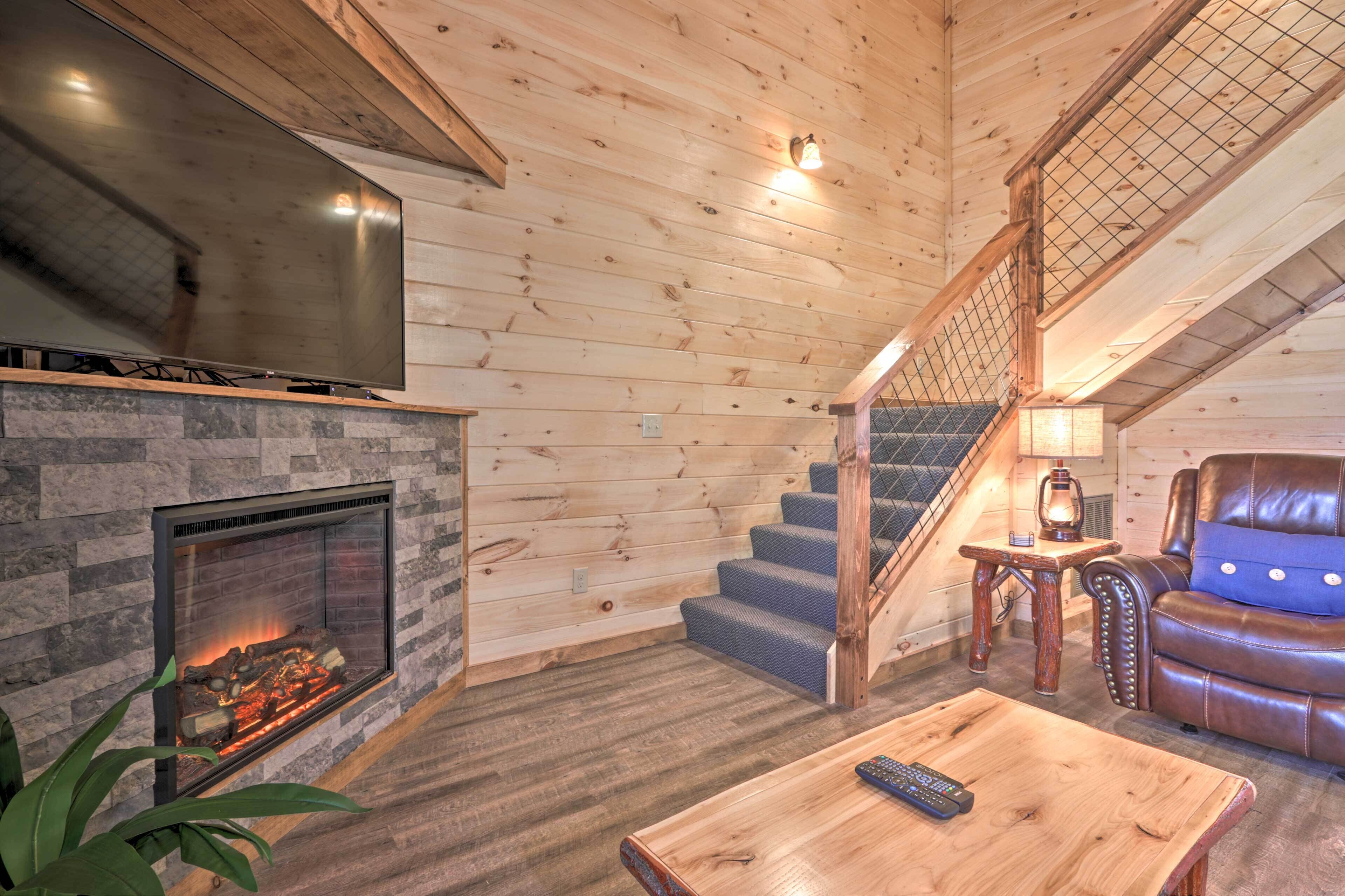 A cozy living room with a stone fireplace, a large flat-screen TV, a wooden staircase, and a leather sofa.