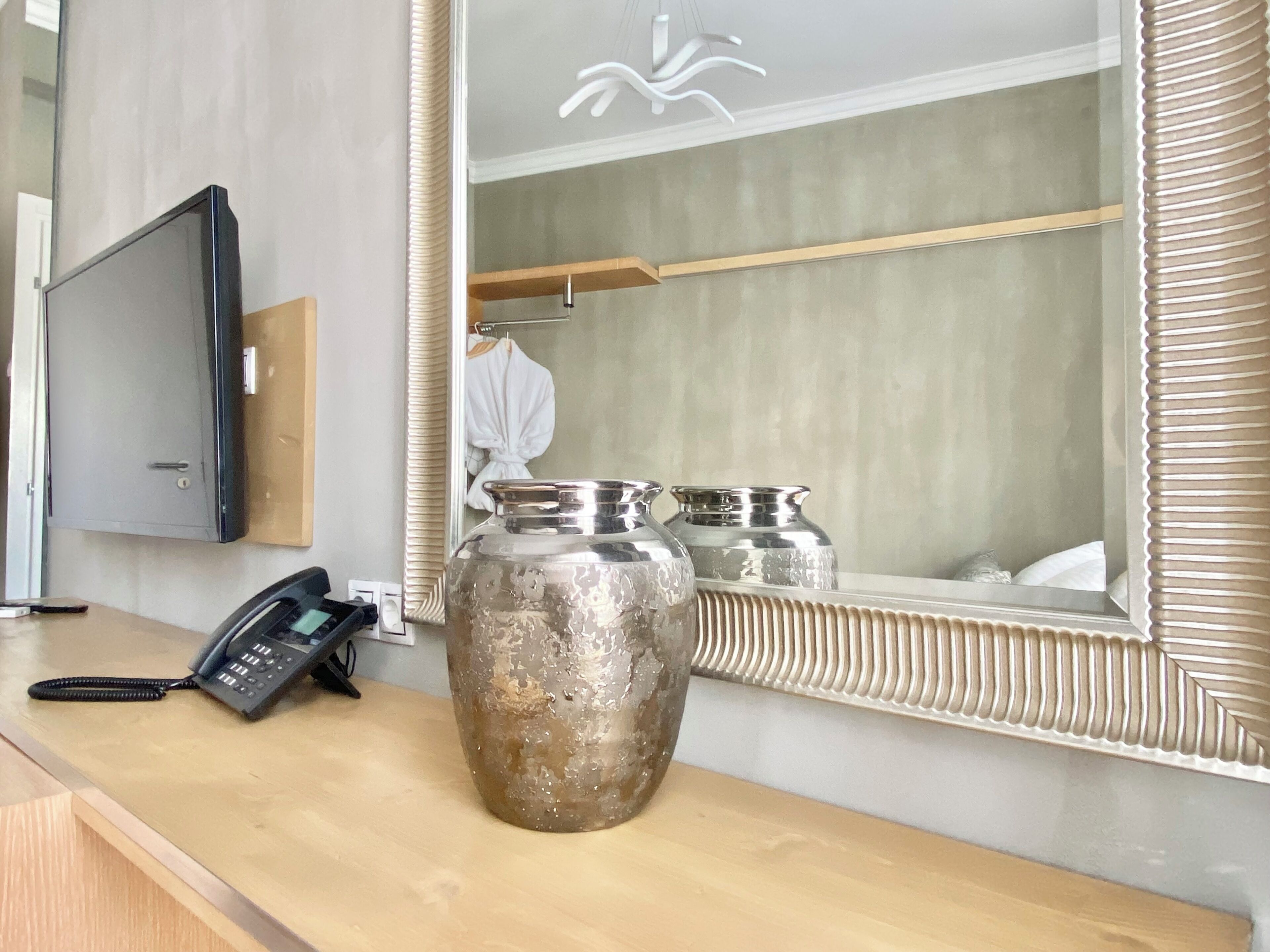 A wooden desk with a telephone, a large mirror with a decorative frame, and two metallic vases.