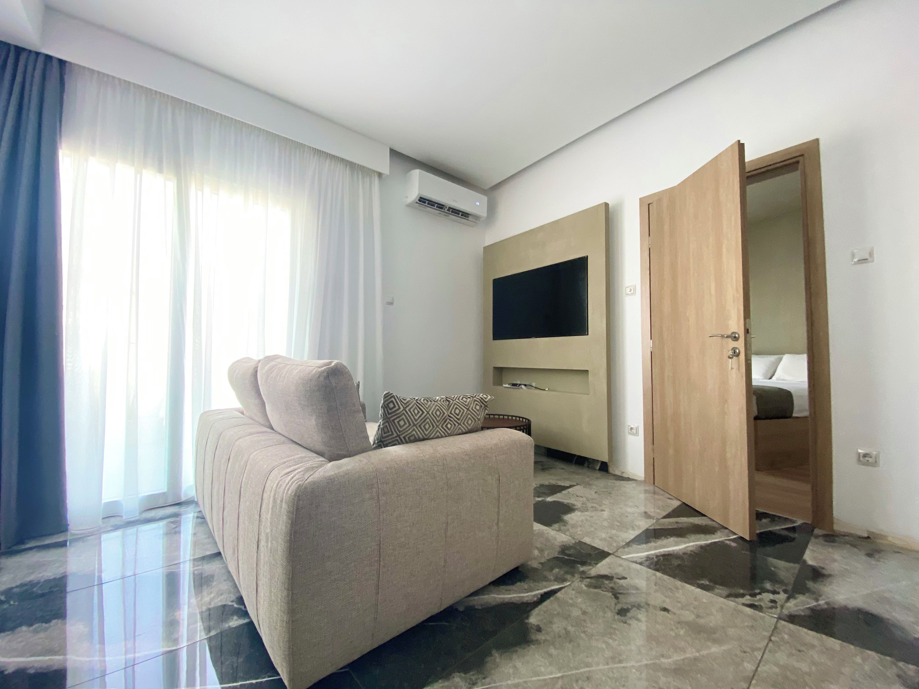 A modern living room with a sofa, a flat-screen TV mounted on the wall, and a door leading to a bedroom.