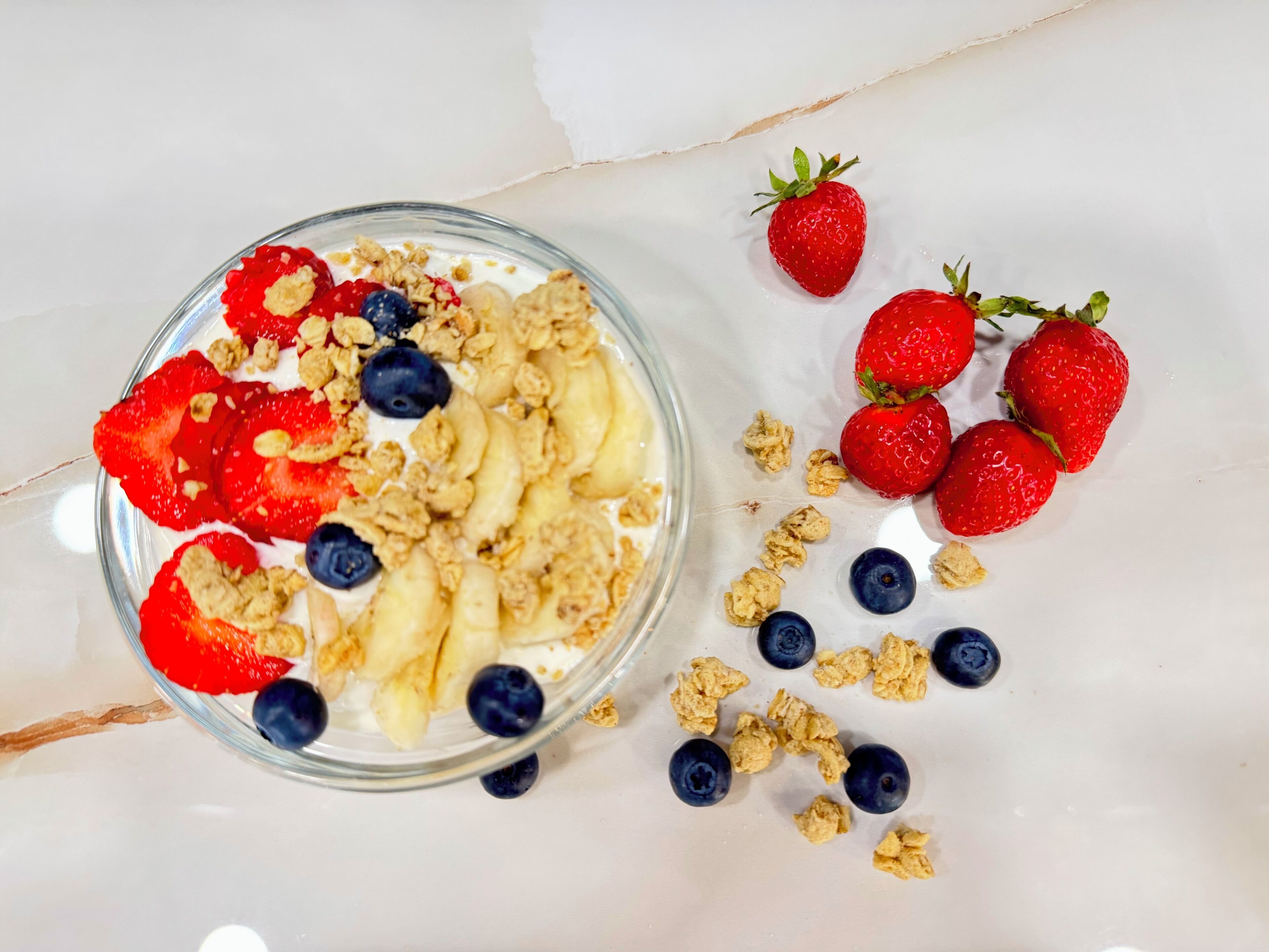 A bowl of yogurt with strawberries, blueberries, and granola on a marble surface.