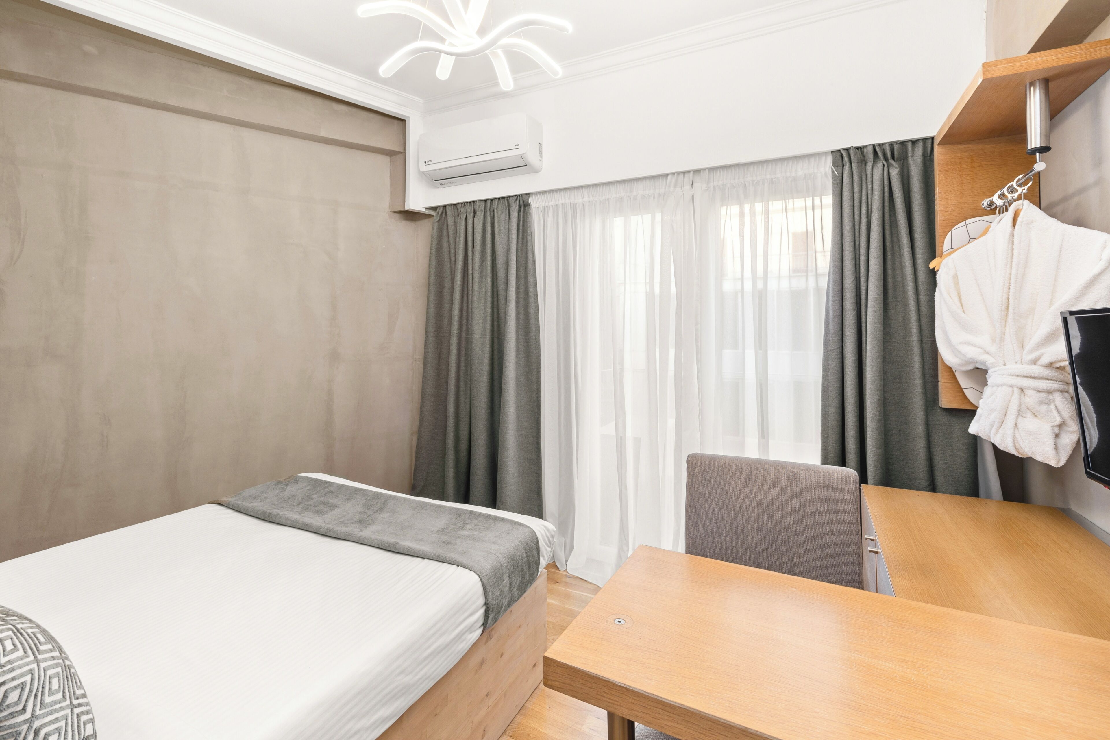 A hotel room with a bed, a desk, a chair, a robe hanging on a rack, and an air conditioning unit.
