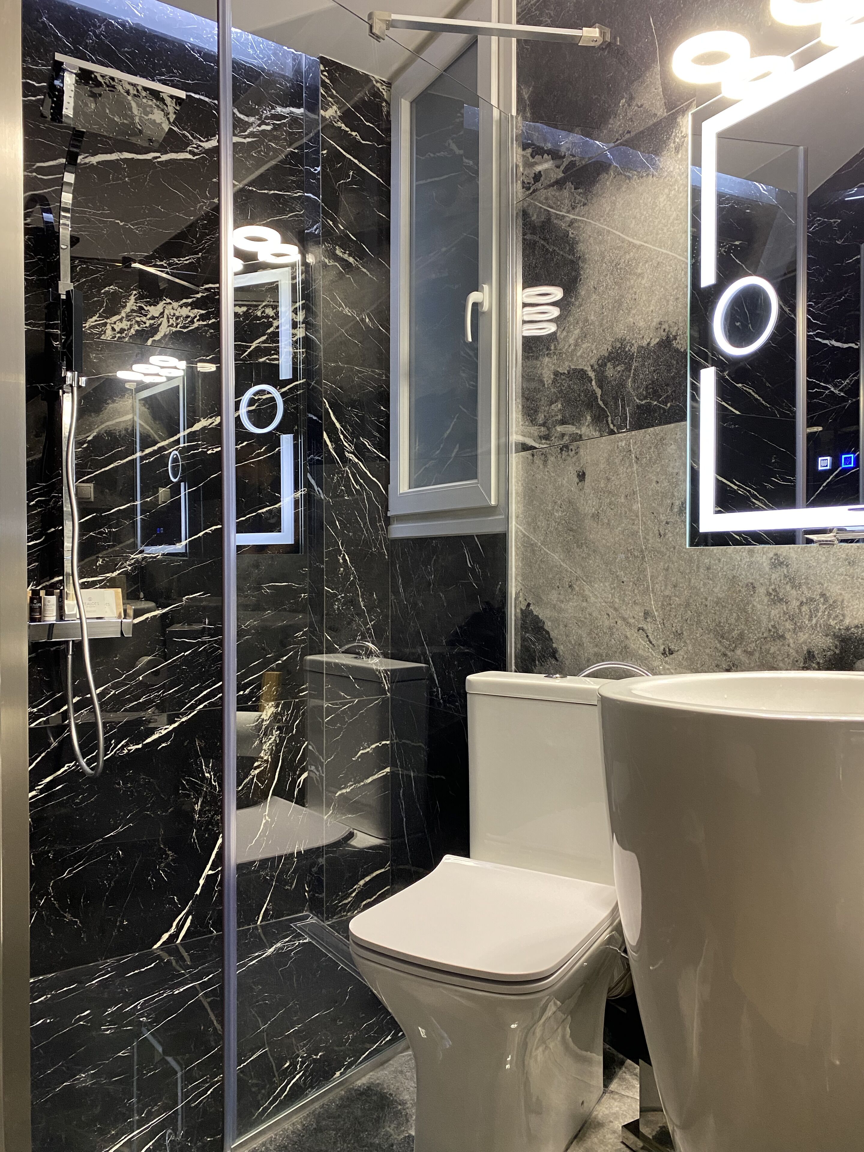 Un baño moderno con salpicadero de mármol, ducha con mampara de vidrio, inodoro y un lavamanos grande con espejo.