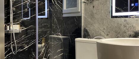 Un baño moderno con salpicadero de mármol, ducha con mampara de vidrio, inodoro y un lavamanos grande con espejo.