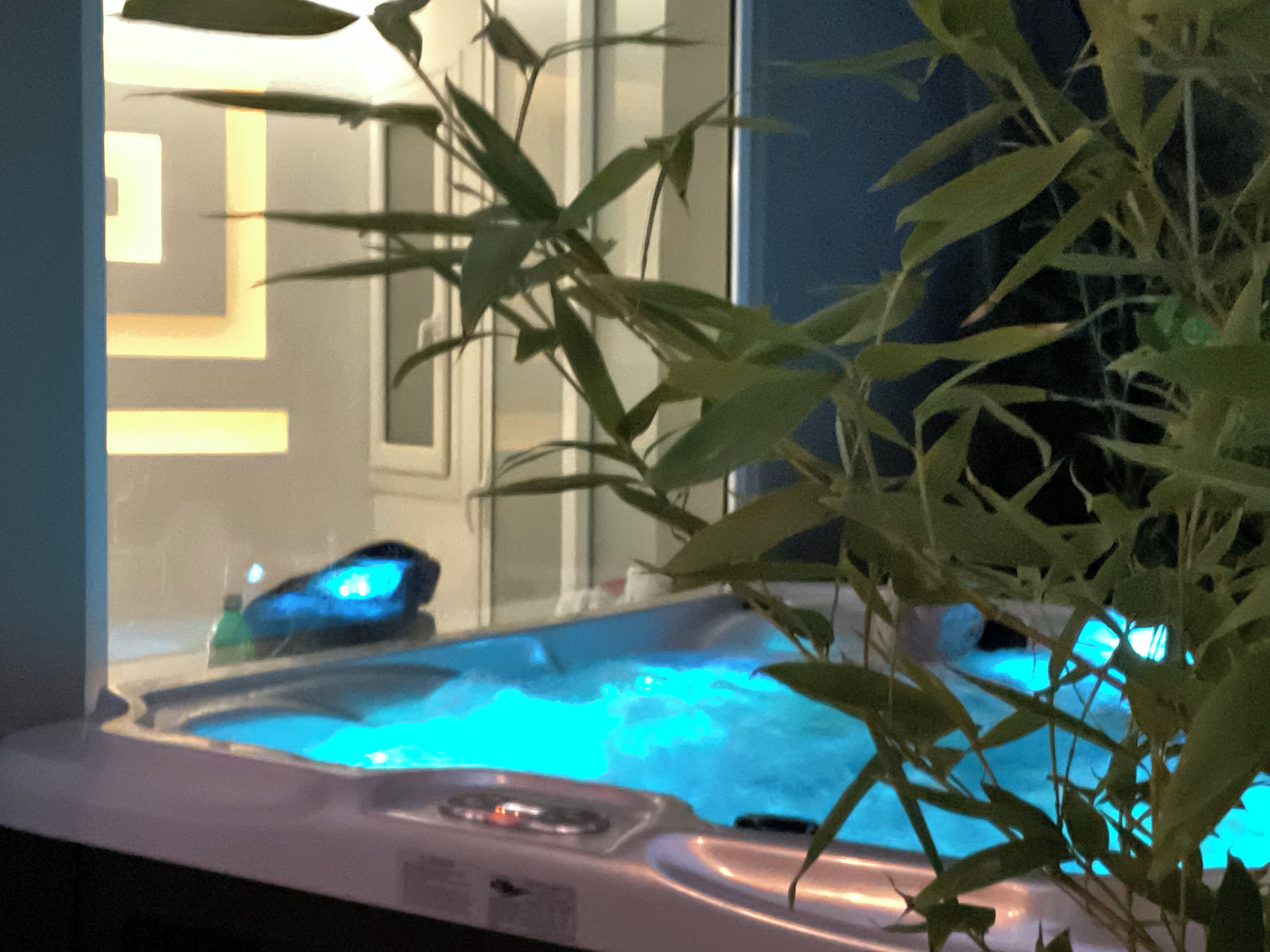 Un jacuzzi con luz azul, una ventana con diseño geométrico y una planta al frente.