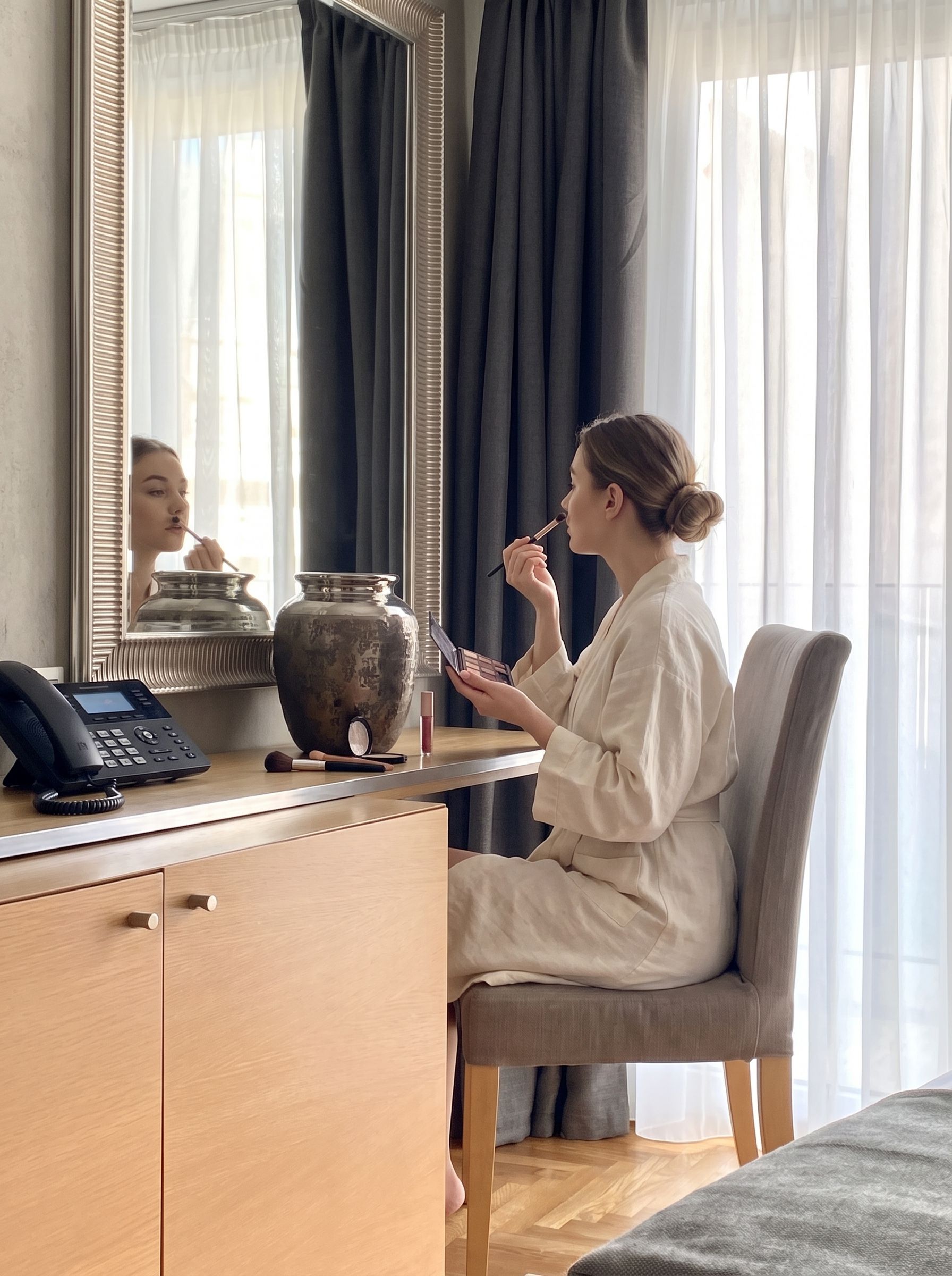 Une personne vêtue d’une robe, assise à un bureau devant lequel se trouvent un miroir, un vase et un téléphone.