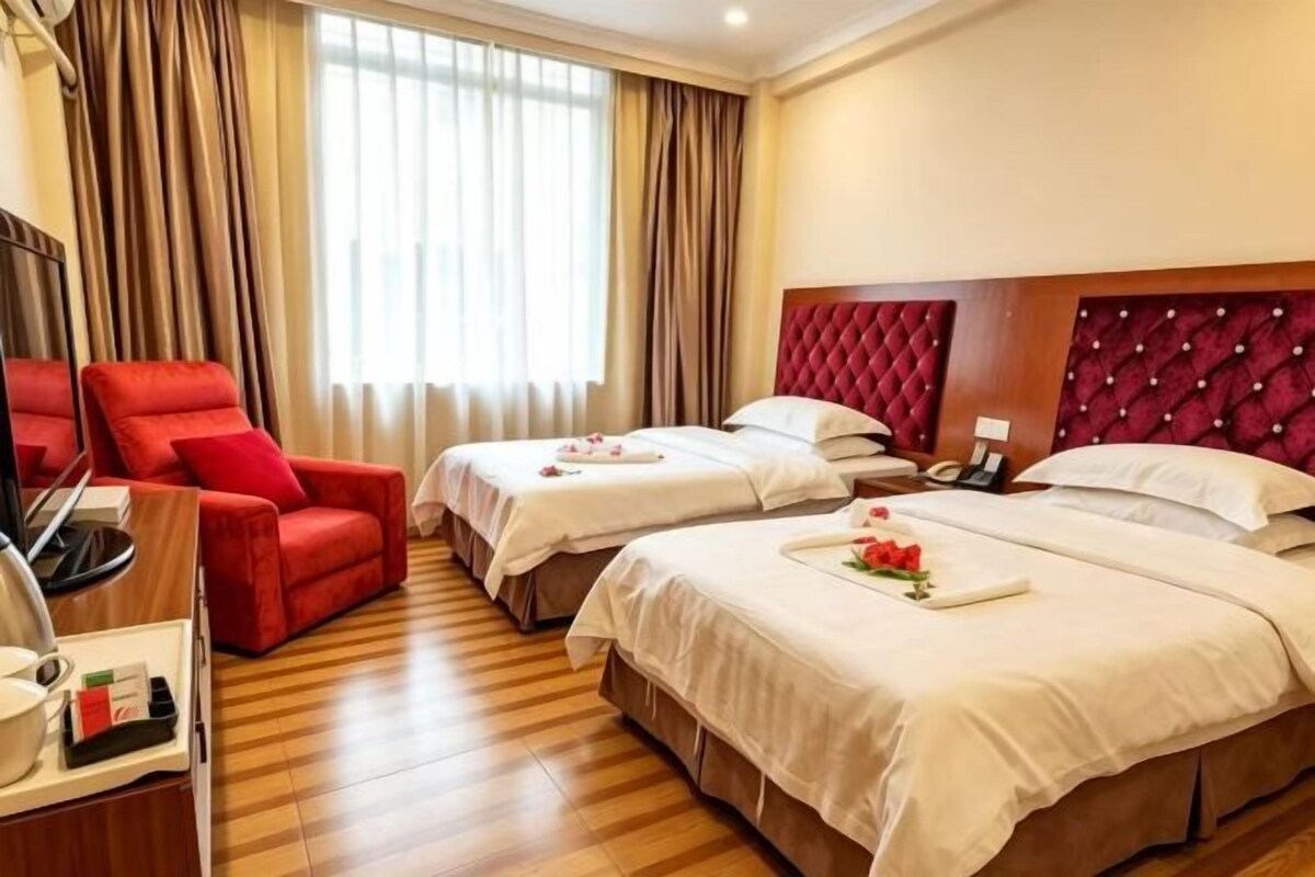 Ein Hotelzimmer mit zwei Betten, einem roten Sessel, einem Schreibtisch mit Lampe und einem großen Fenster mit Vorhängen.