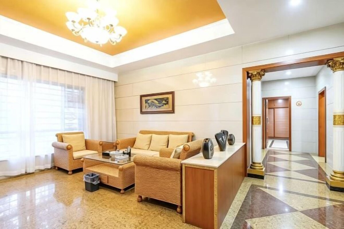 Ein Hotelzimmer mit Sofagarnitur, einem Couchtisch und einem kleinen Tisch mit einer Vase. Ein Fenster mit Vorhängen, eine Kristallleuchter und ein gerahmtes Bild sind an der Wand zu sehen.