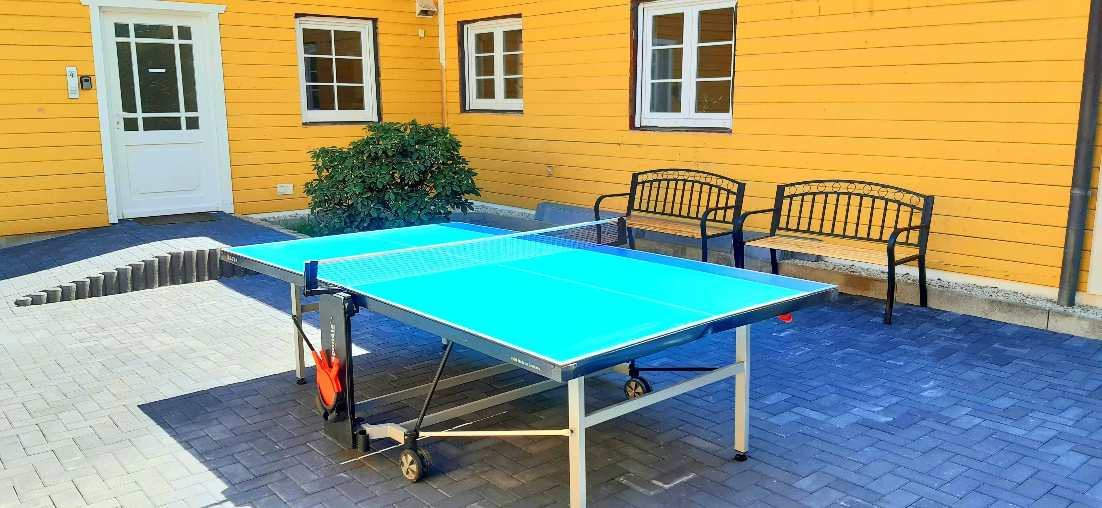 An outdoor ping pong table with chairs, a small bush, and a yellow building with windows.