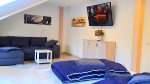 Un salon moderne avec un canapé, une petite table blanche, un téléviseur fixé au mur et un meuble en bois orné d’une plante.